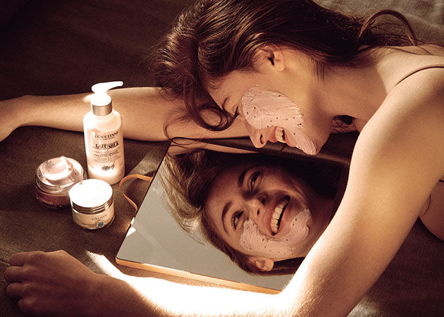 Woman looking into mirror applying soothing face mask, L'OCCITANE Beauty Tips, L'OCCITANE  Cleansing Milk Facial Make-up Remover, L'OCCITANE Soothing Face Mask, L'OCCITANE Shea Light Comforting Face Cream  
