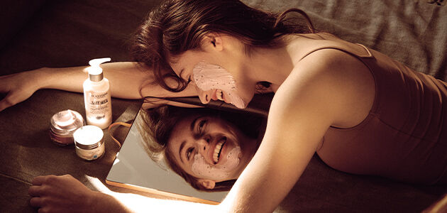Woman looking in mirror in order to apply L'OCCITANE Soothing Face Mask to face.