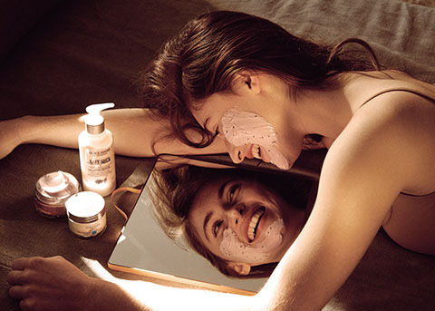 Woman looking into mirror applying soothing face mask, L'OCCITANE Beauty Tips, L'OCCITANE  Cleansing Milk Facial Make-up Remover, L'OCCITANE Soothing Face Mask, L'OCCITANE Shea Light Comforting Face Cream  