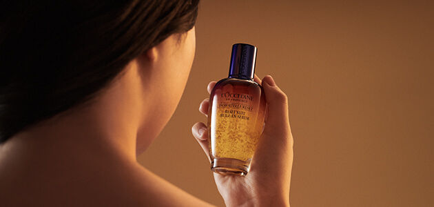 The image shows a woman looking directly at the L'OCCITANE Immortelle Reset Serum